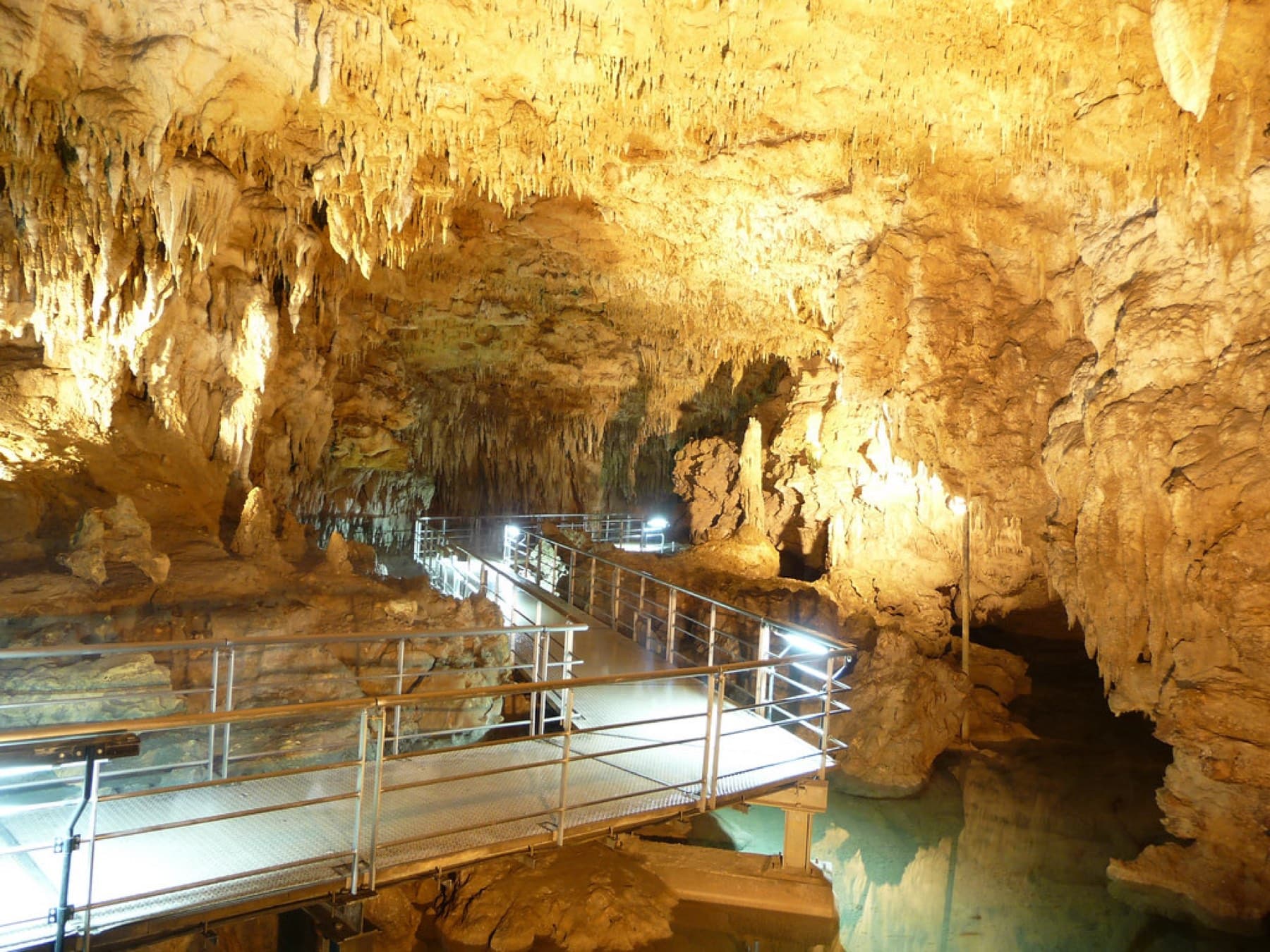 A steel walkway crosses clear water beneath a ceiling dense with pale stalactites in the Gyokusendo limestone cave