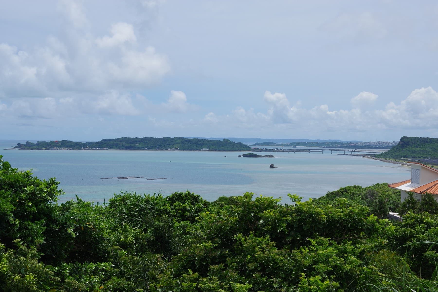 Green scrub frames a wide view south from Kafu Banta over a calm bay to the wooded island of Hamahiga and the long low line of its bridge