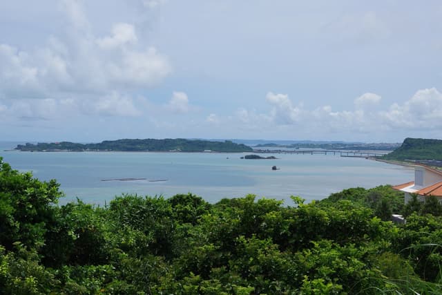 Green scrub frames a wide view south from Kafu Banta over a calm bay to the wooded island of Hamahiga and the long low line of its bridge