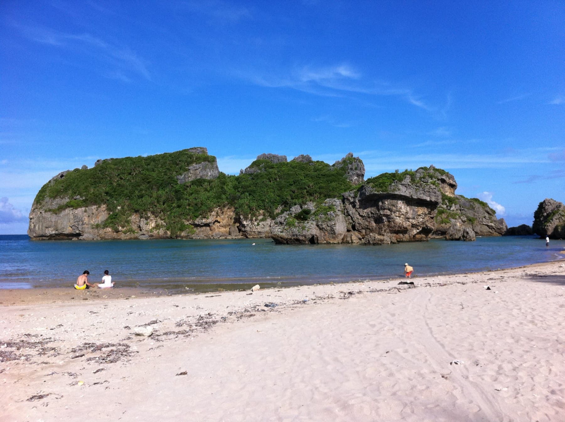 A few swimmers rest on the white sand of a beach on Hamahiga facing the rocky green islet of Kuba across calm water