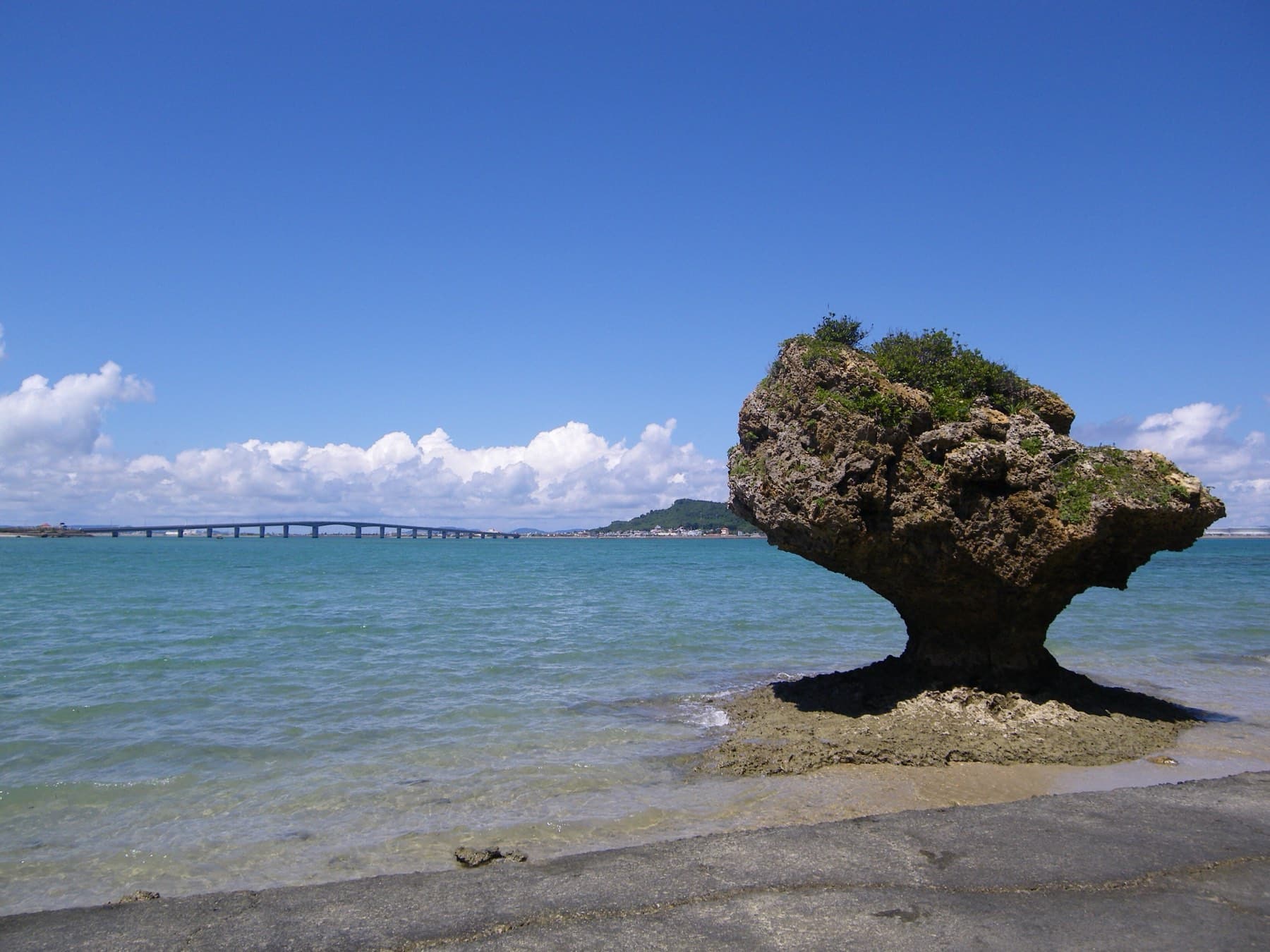 A mushroom-shaped rock crowned with green shrubs stands in shallow water off Hamahiga, and the long Hamahiga Bridge crosses the bay behind it under a blue sky