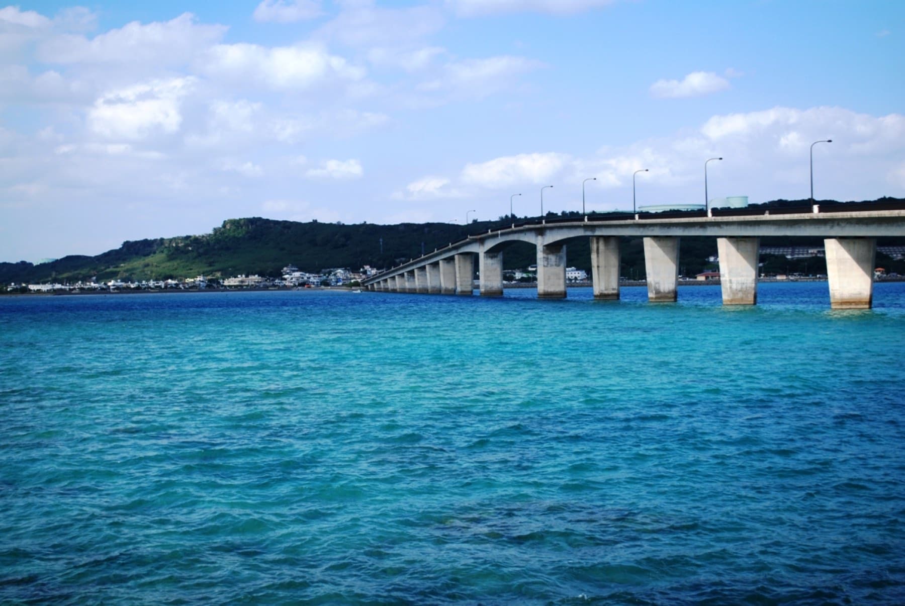 The concrete piers of the Hamahiga Bridge march across deep blue water toward the wooded ridge and white village of Hamahiga