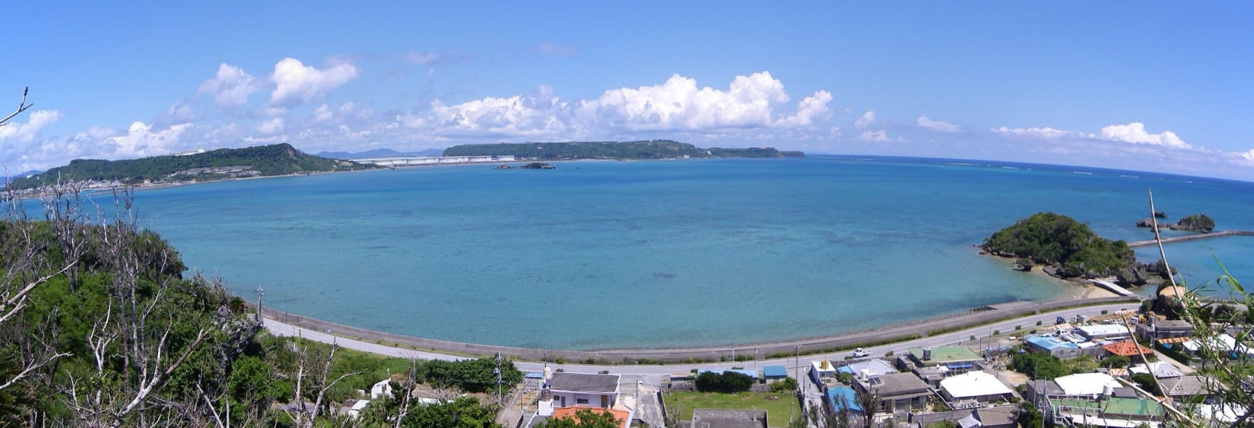 A wide view from a lookout on Hamahiga takes in a calm blue bay, a curving seawall above village roofs, and the long green islands of Henza and Miyagi