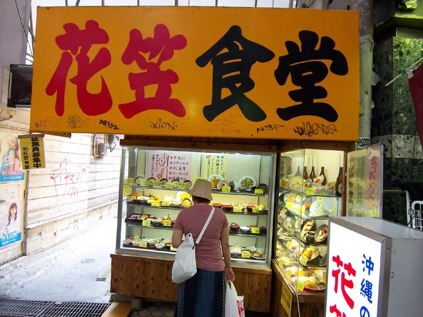 A woman studies the plastic food display under the big orange Hanagasa Shokudo sign in a covered market lane in Makishi