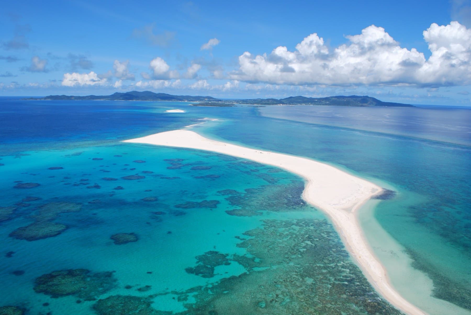 The white sandbar of Hatenohama curves for kilometres through turquoise shallows dotted with dark coral heads, and Kume-jima rises green on the horizon