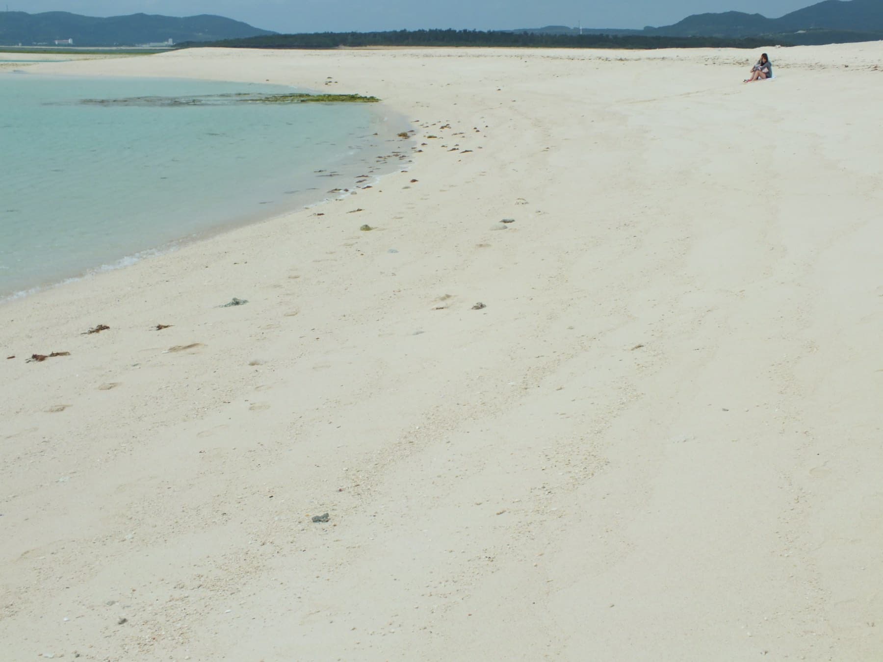 A tide pool lagoon curves along the white sand of Hatenohama, with green weed in the shallows and the hills of Kume-jima low on the horizon