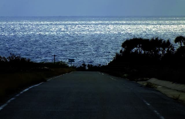 A narrow road on Hateruma runs straight between dark scrub toward a sheet of glittering sea, lit silver by low winter sun through cloud.