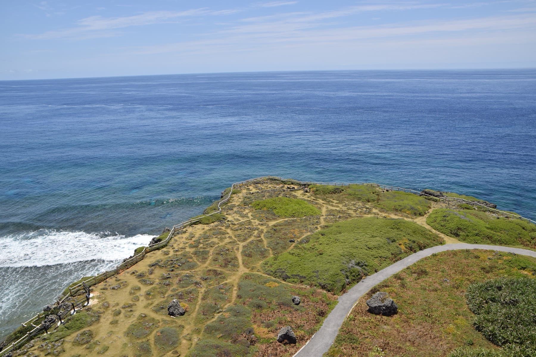 A paved path winds over the grassy, rock-studded spine of Higashi-Hennazaki as white surf breaks against its flank and the Pacific stretches deep blue beyond.