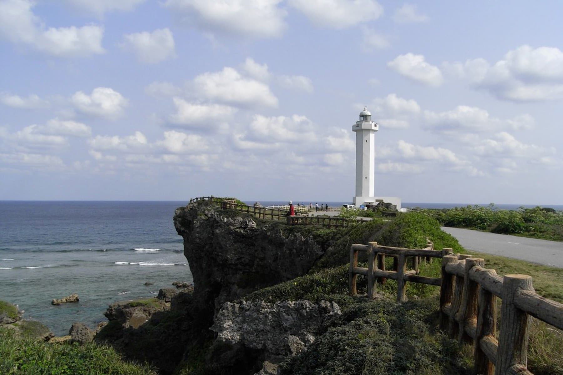 A wooden fence follows the edge of a dark limestone cliff toward the white lighthouse at Higashi-Hennazaki, where the reef breaks offshore.