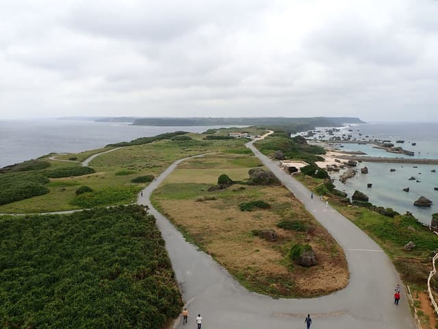 Seen from the lighthouse, the road on Higashi-hennazaki loops back along the grassy cape between low scrub, with walkers on the tarmac and boulders in the shallows.