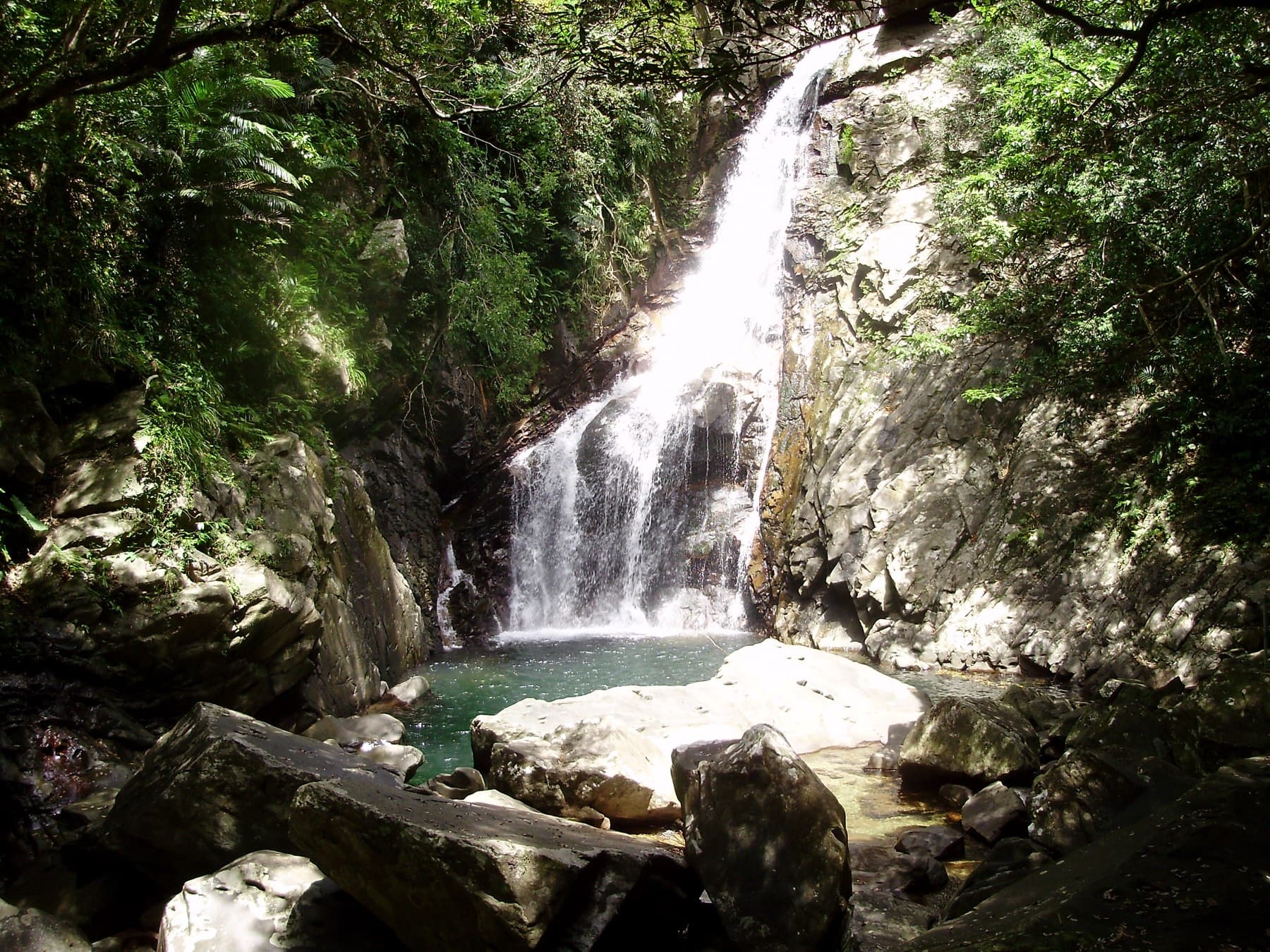 Grey boulders ring the green plunge pool at the foot of Hiji Falls, where the water fans out over a ledge in the forested gorge
