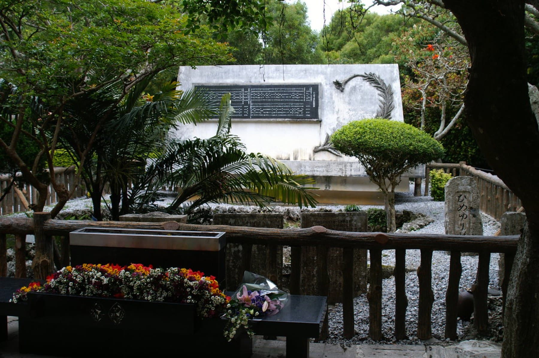Flowers and incense sit before the Himeyuri monument in Itoman, a white stone wall carved with names behind a wooden railing in the shade of trees