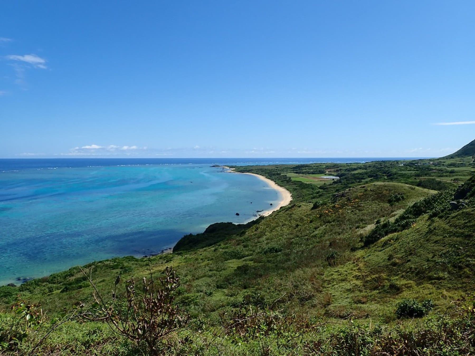 Green slopes on the Hirakubo peninsula fall to a pale sand cove and a wide shelf of reef water along the far north coast of Ishigaki.