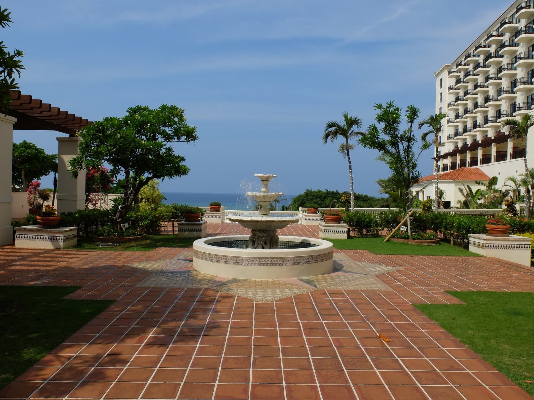 A white fountain stands in a terracotta-tiled patio at Hotel Nikko Alivila in Yomitan, with palms, the hotel's arched wings and a glimpse of the sea beyond