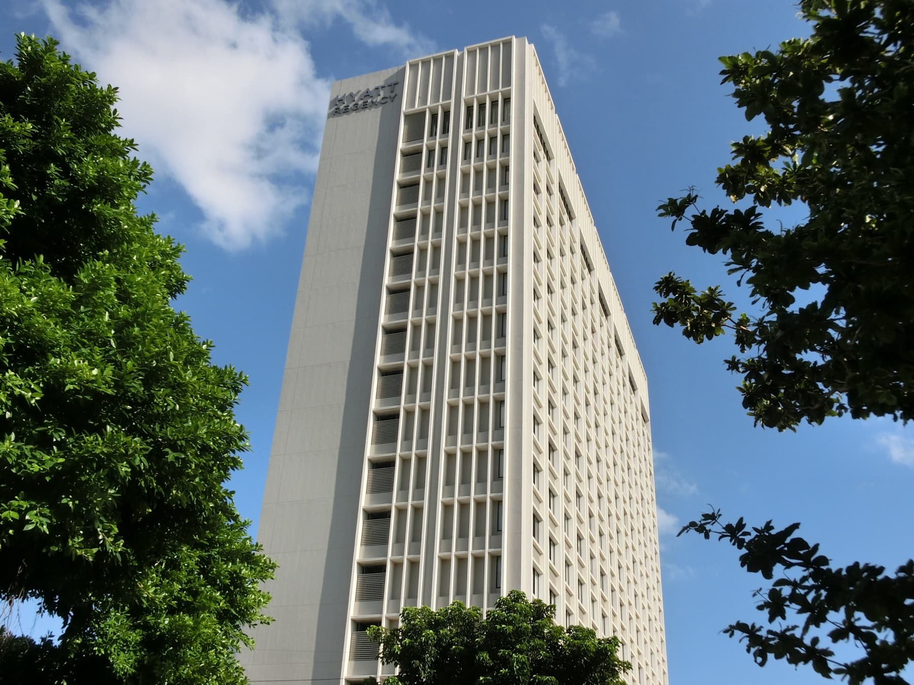 The tall pale tower of the Hyatt Regency Naha rises between green trees into a blue sky streaked with white cloud