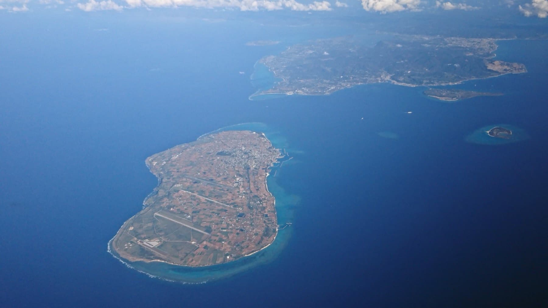 Ie-jima lies flat and oval in deep blue sea, ringed by a pale band of reef, with the Motobu Peninsula and Minna faint beyond it