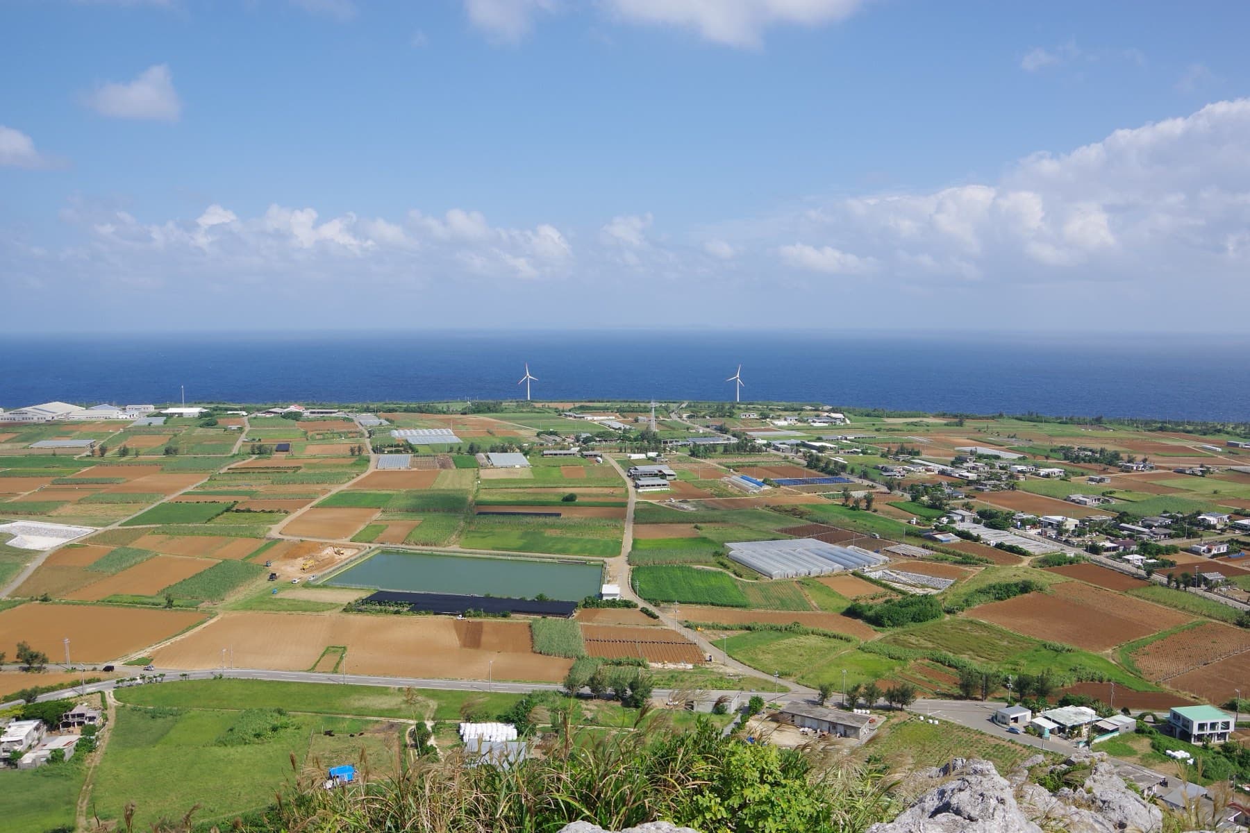 From the top of Gusuku-yama, flat farmland on Ie-jima spreads out in a patchwork of red earth and green fields cut by straight farm roads, with wind turbines on the far coast.