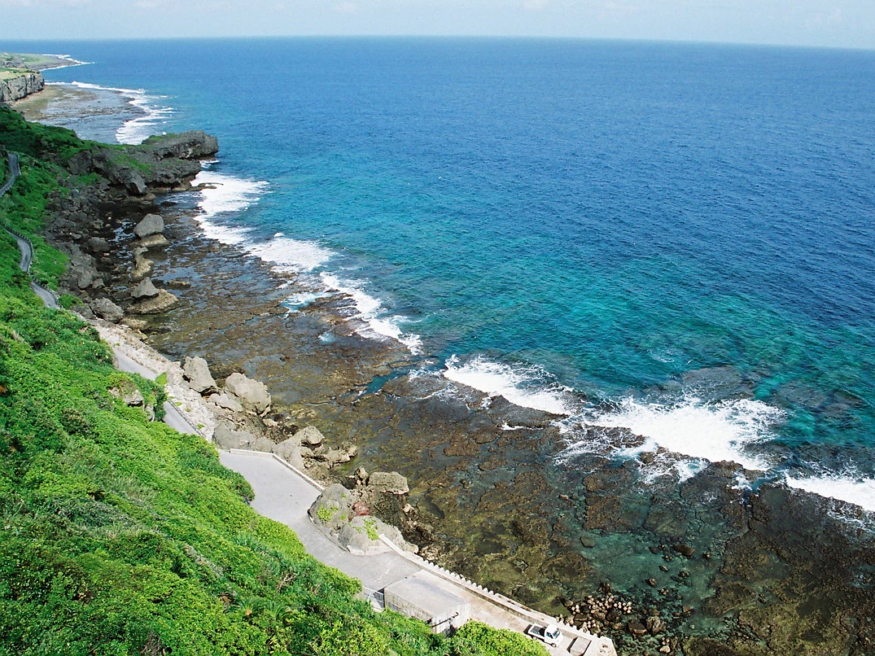 A narrow road runs along the foot of green cliffs at Waji on Ie-jima, beside a rocky reef where waves break white on deep blue water.