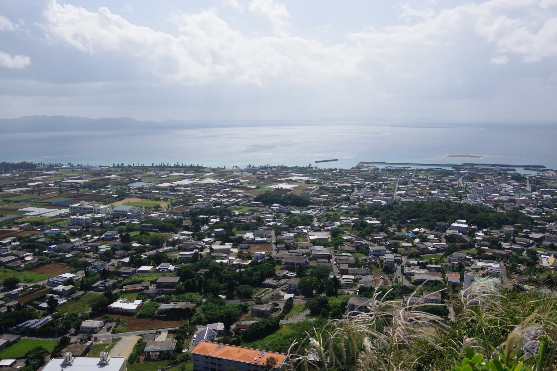 The rooftops of Ie village fill the south side of Ie-jima below Gusuku-yama, and the port breakwater reaches into a hazy sea toward Motobu