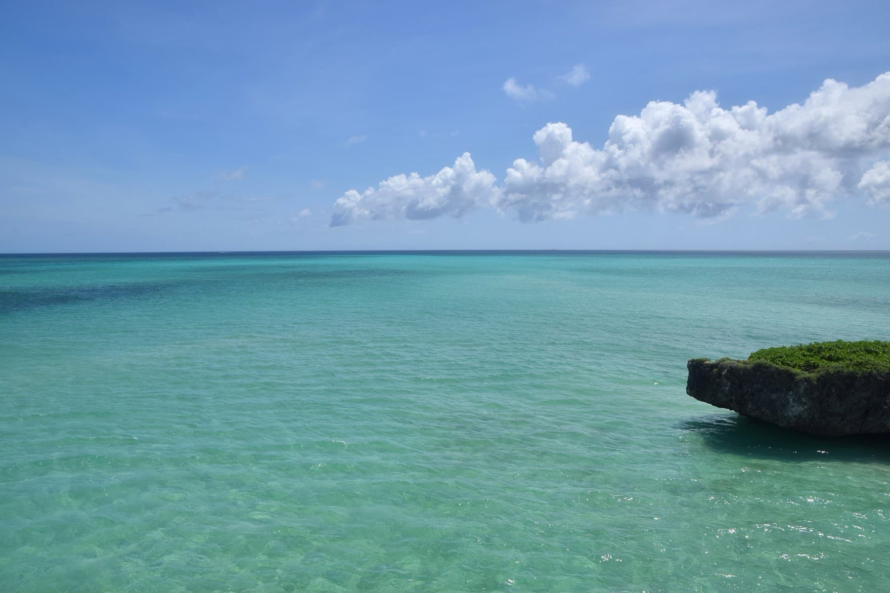 Emerald water over a sandy reef stretches toward the horizon beside a rocky ledge near the Ikema Bridge on a bright summer morning.