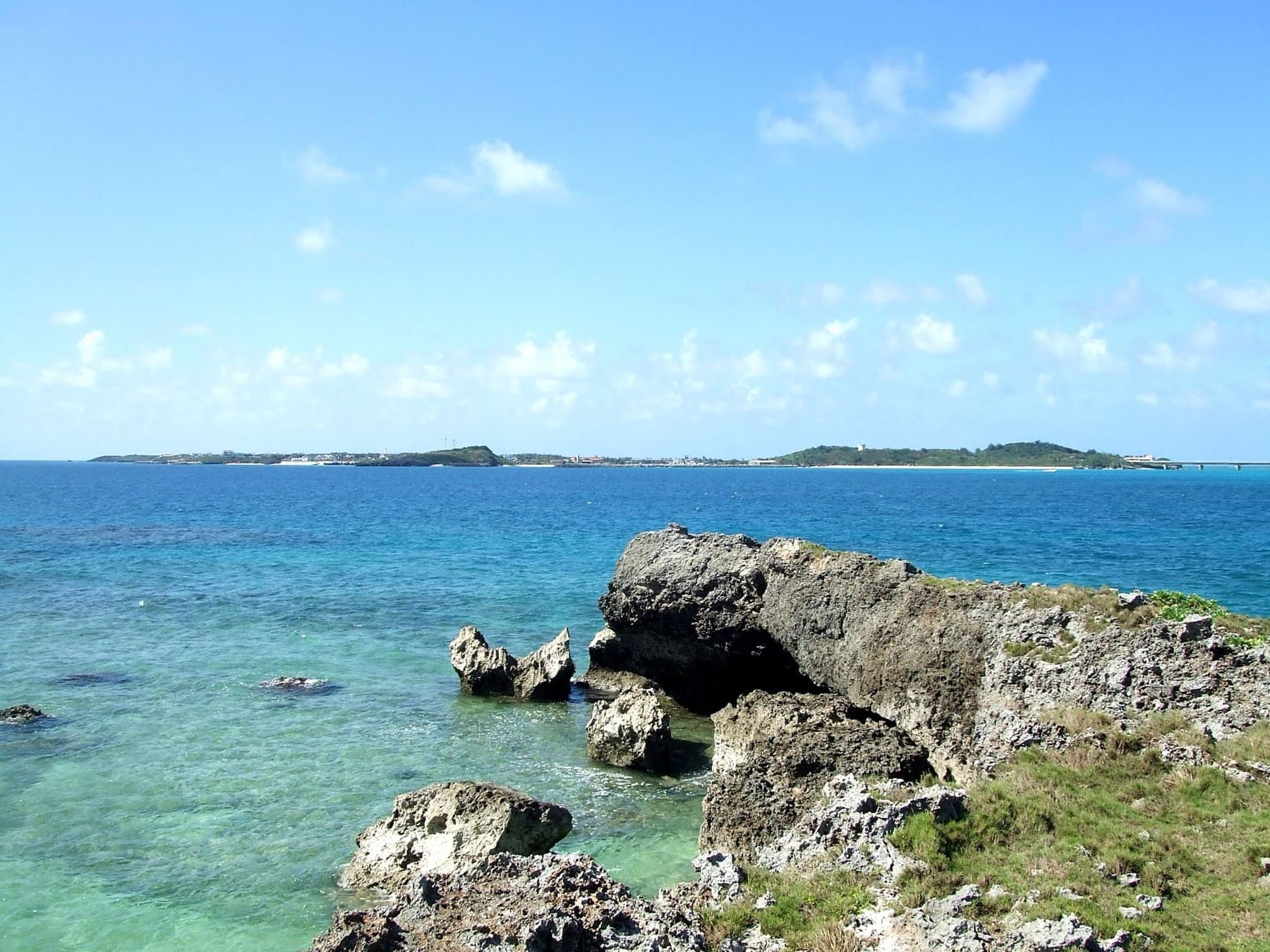 Low and flat, Ikema lies along the horizon beyond clear reef water and a ledge of rough coral rock on a bright summer day.
