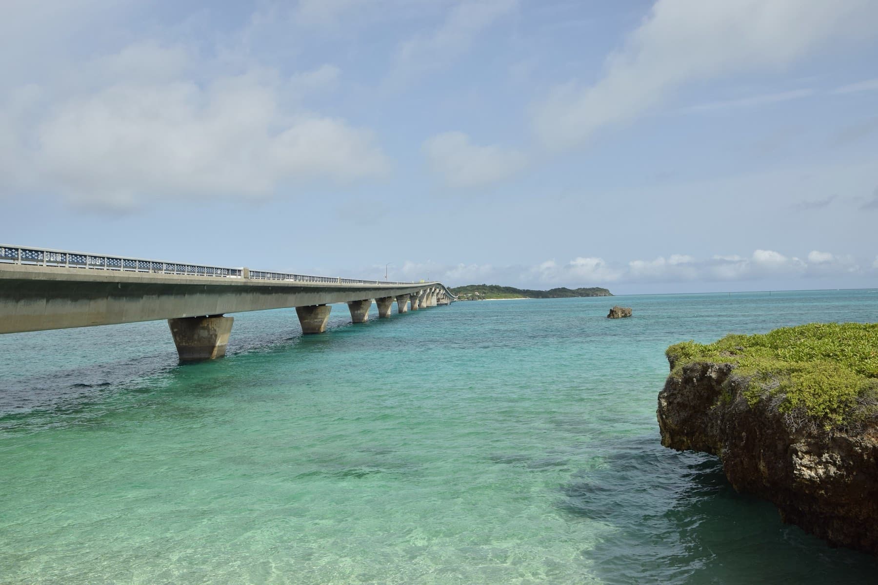 Morning light shows the sandy bottom through clear shallow water beside the long, low Ikema Bridge and a rocky limestone outcrop.