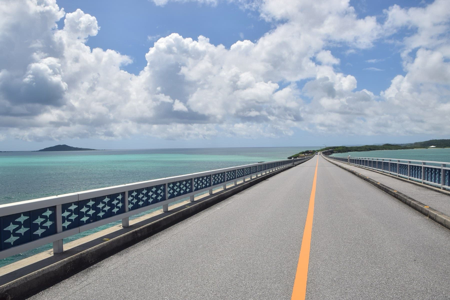 An orange centre line leads across the deck of the Ikema Bridge between patterned railings as summer clouds pile up over the sea.