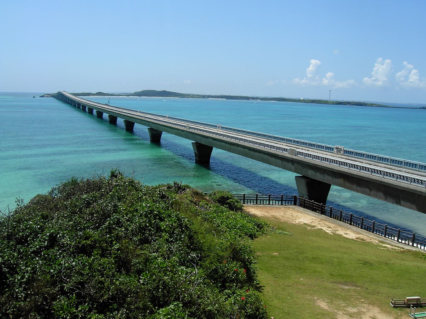 The Ikema Bridge strides on concrete piers across green and turquoise reef water from a grassy headland on Miyako toward the low island of Ikema.