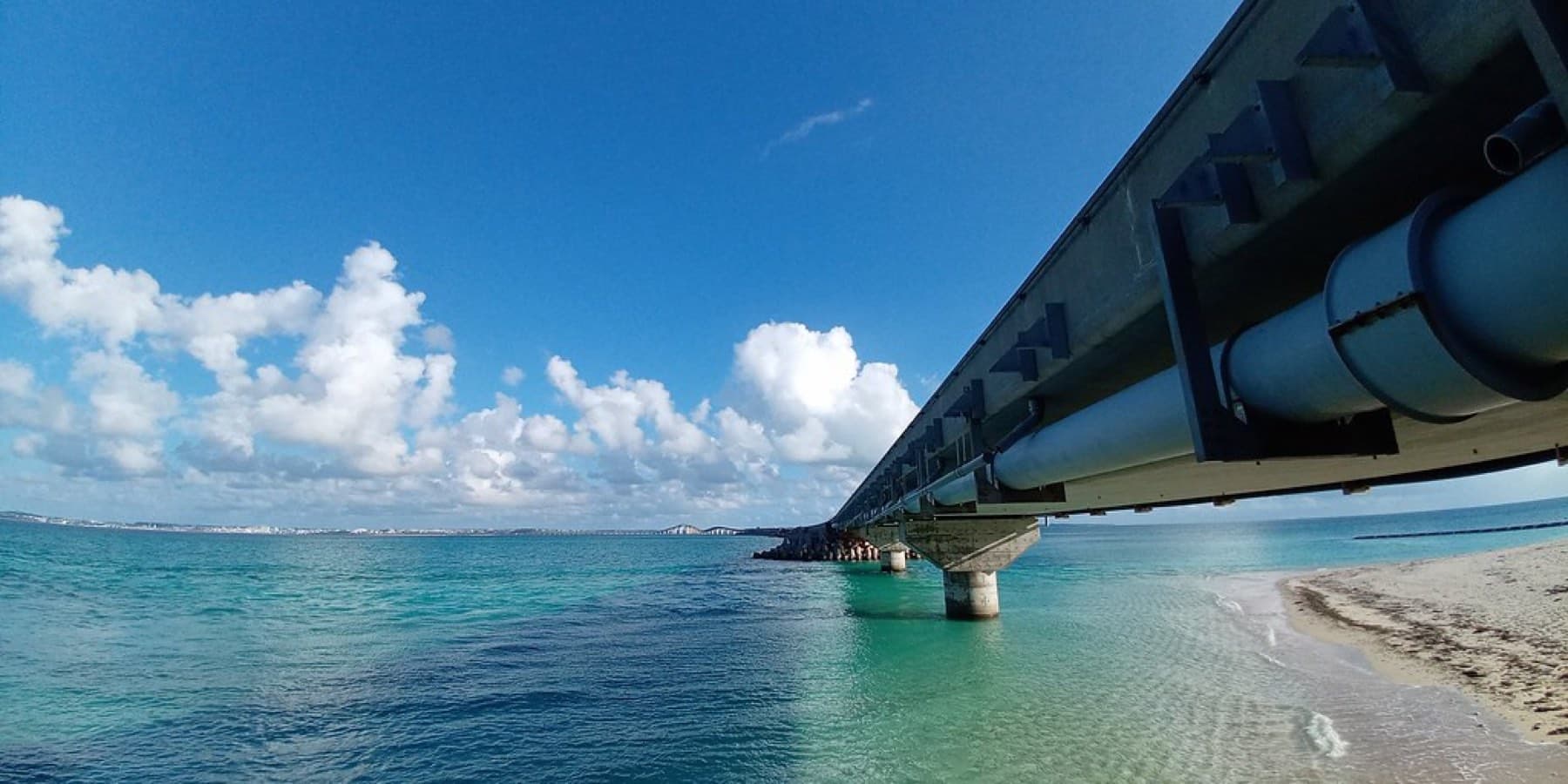 The underside of the Irabu Bridge frames clear water and puffy clouds from a small beach beside its approach.