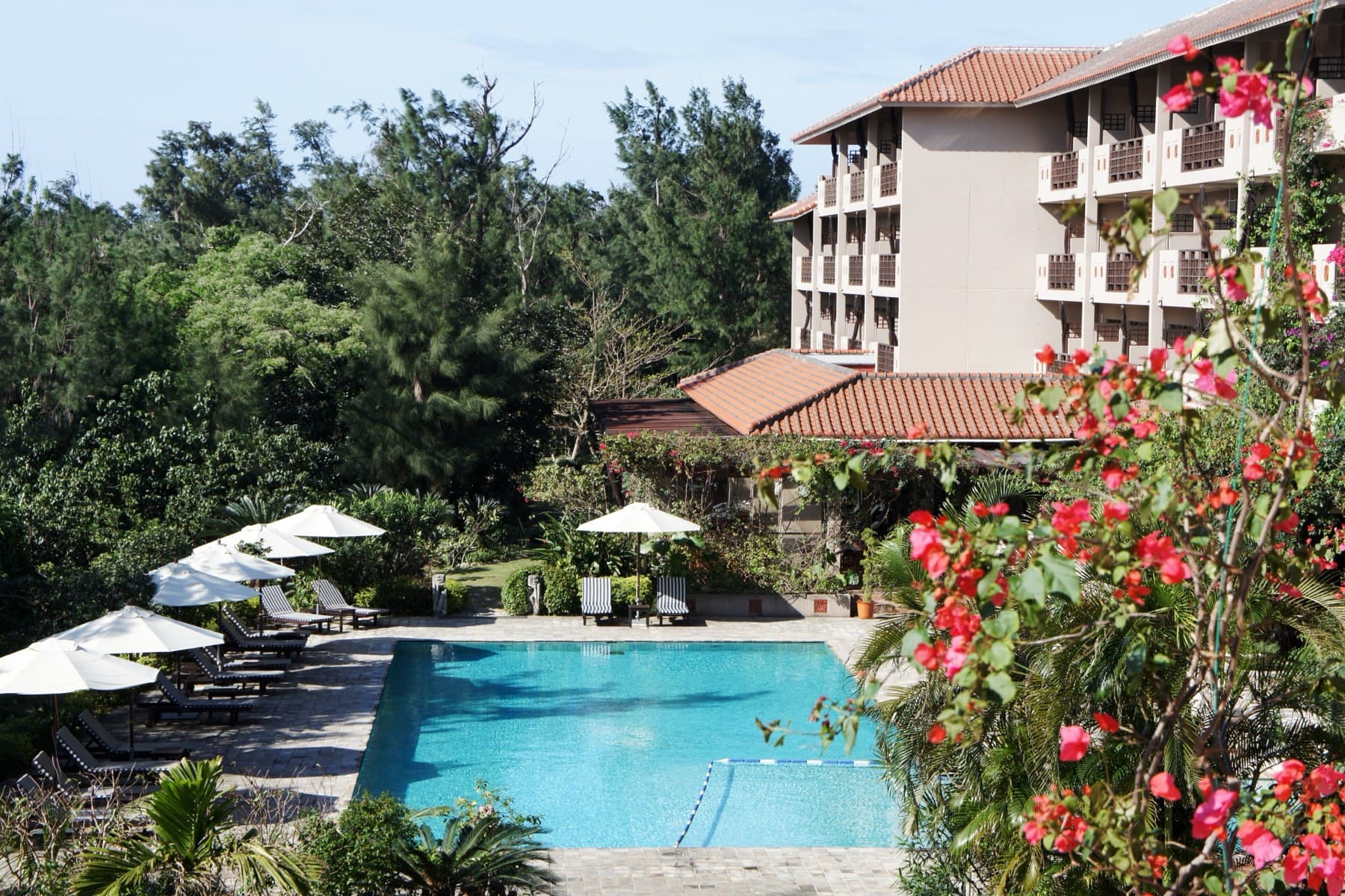 Red flowers frame the pool and white umbrellas in the garden of the Iriomote Hotel by Hoshino Resorts behind Tsukigahama.