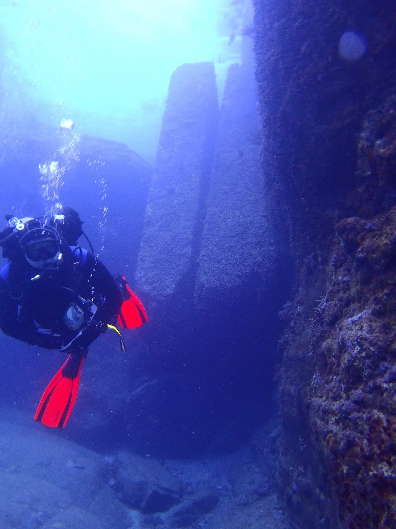 A diver in red fins swims along the sheer stone face of the Yonaguni Monument in deep blue water.