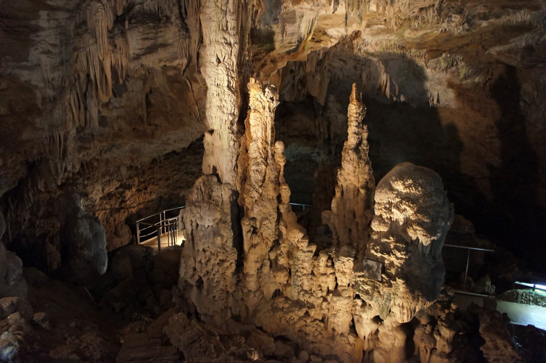 Tall stalagmites crowd a lit chamber of the Ishigaki-jima Limestone Cave beside the railed walkway that threads through it.