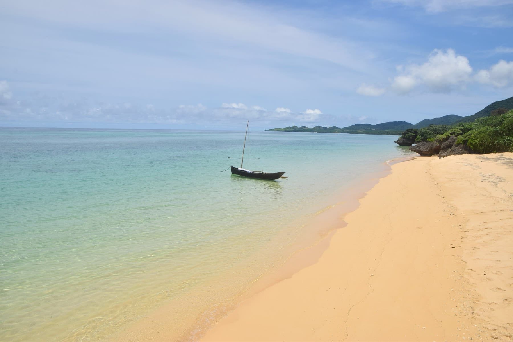 A small boat rides at anchor off the golden sand of Ishigaki Sunset Beach as the shore curves away toward a green headland.