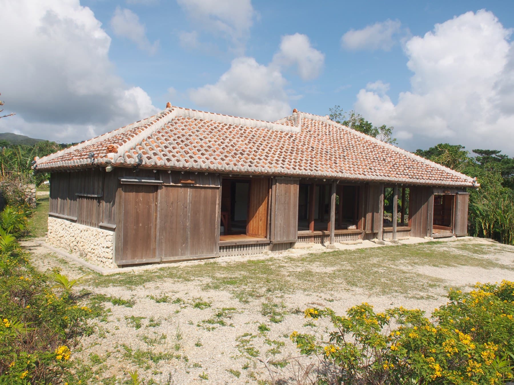 A low wooden house with a hipped roof of red Ryukyu tiles sits among yellow flowers at Ishigaki Yaima-mura under a summer sky.