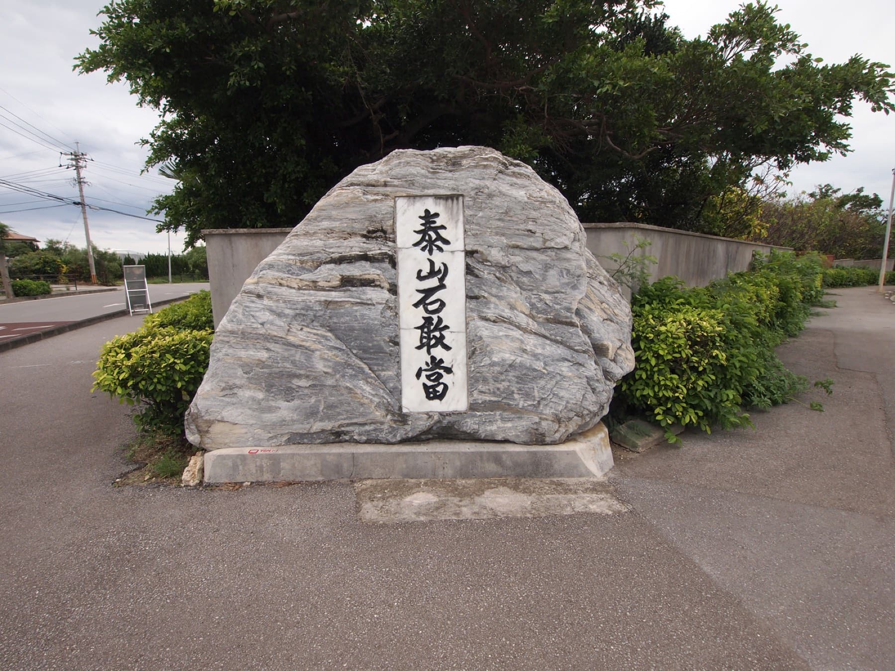 A large grey boulder carved with the words Taishan Ishiganto stands on a low plinth at a road junction on Ishigaki, backed by hedges and trees