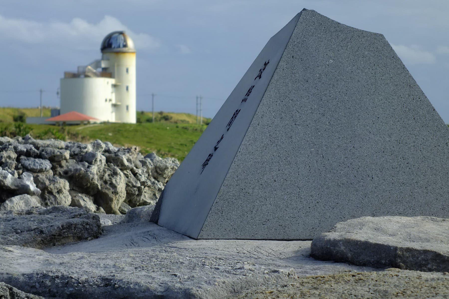 A grey stone pyramid at Japan's Southernmost Monument stands on the coral rock of Takanasaki with the white Hateruma Star Observatory behind it.
