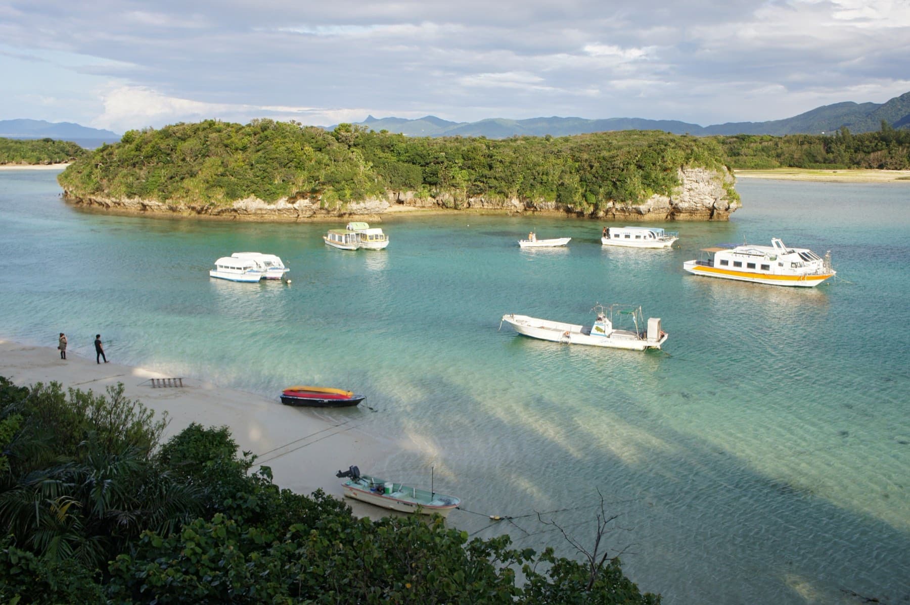Glass-bottom boats ride at anchor over pale green shallows in Kabira Bay, where a wooded limestone islet sits below Ishigaki's hills under soft cloud.