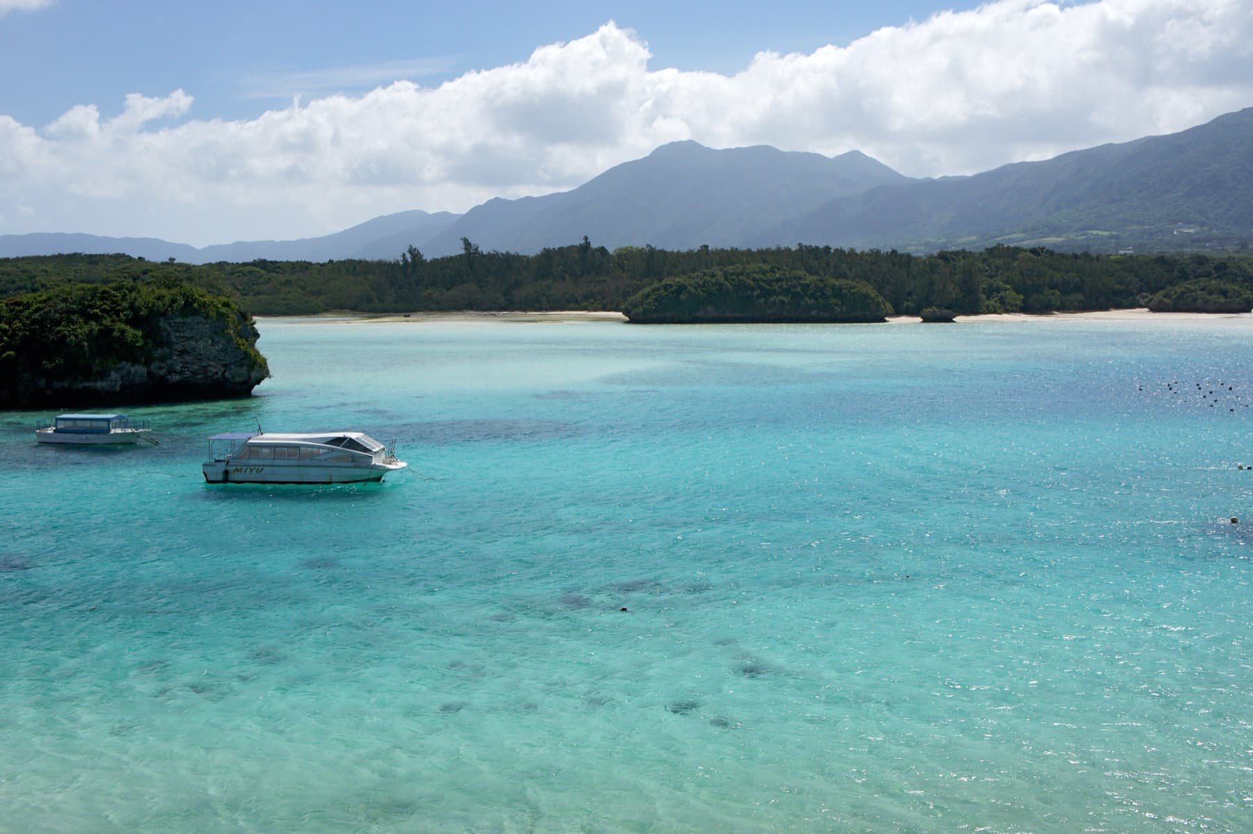 A glass-bottom boat floats over clear shallow water in Kabira Bay while small wooded islets and a range of mountains stand behind it.