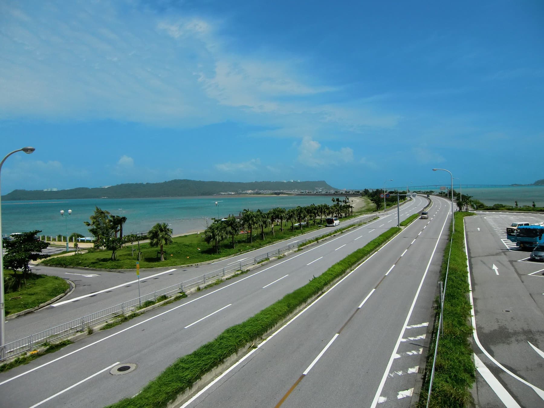Four lanes of the Kaichu Doro causeway run straight across the sea toward Henza, with palms along the shoulder and a long green island on the horizon