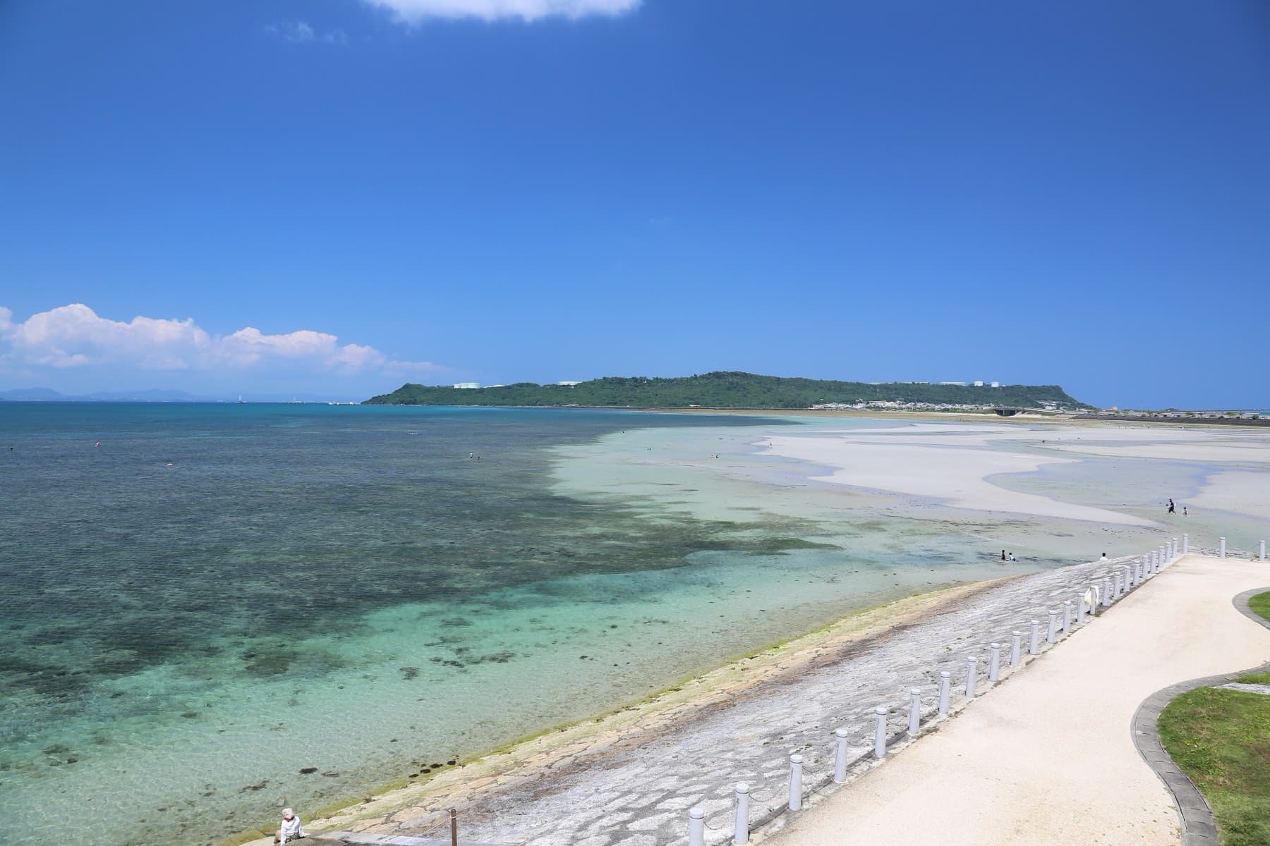 Low tide exposes pale sand flats and reef in clear shallow water beside the Kaichu Doro, with the green ridge of Henza across the bay under a deep blue sky