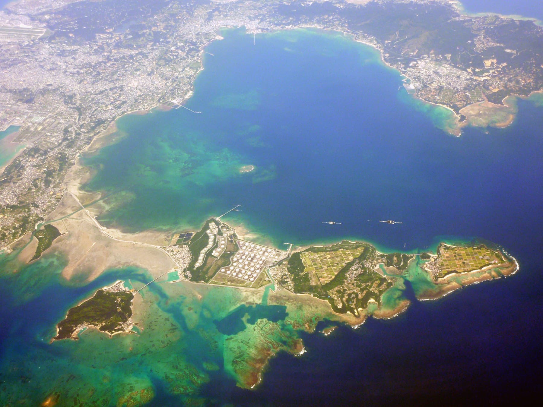An aerial view over Kin Bay follows the Kaichu Doro causeway out to Henza and its oil tanks, and pale green reef surrounds Miyagi and Ikei farther along the chain