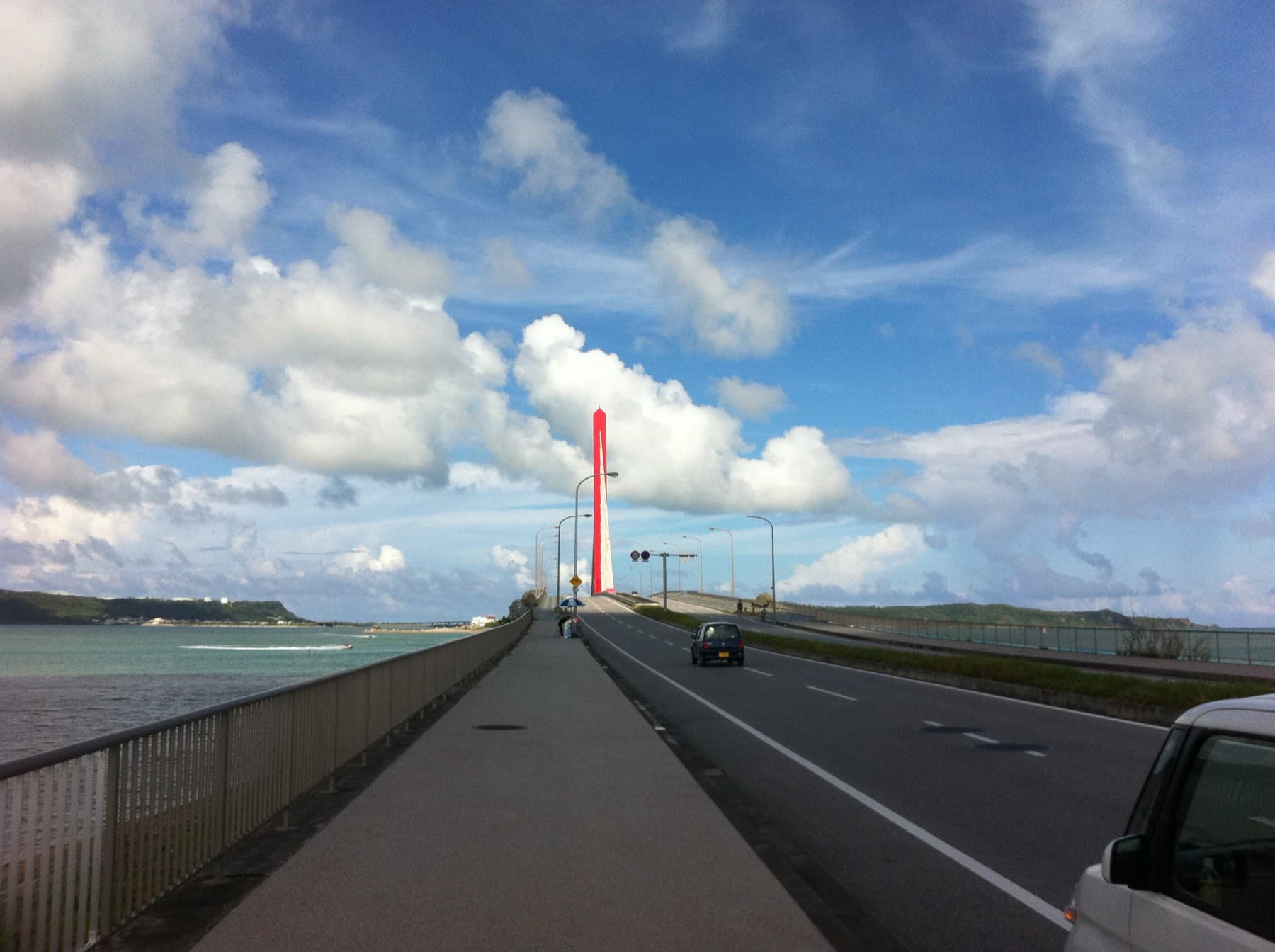 A wide footway runs beside the Kaichu Doro causeway road toward its red bridge tower, with the sea on the left and tall summer clouds overhead.