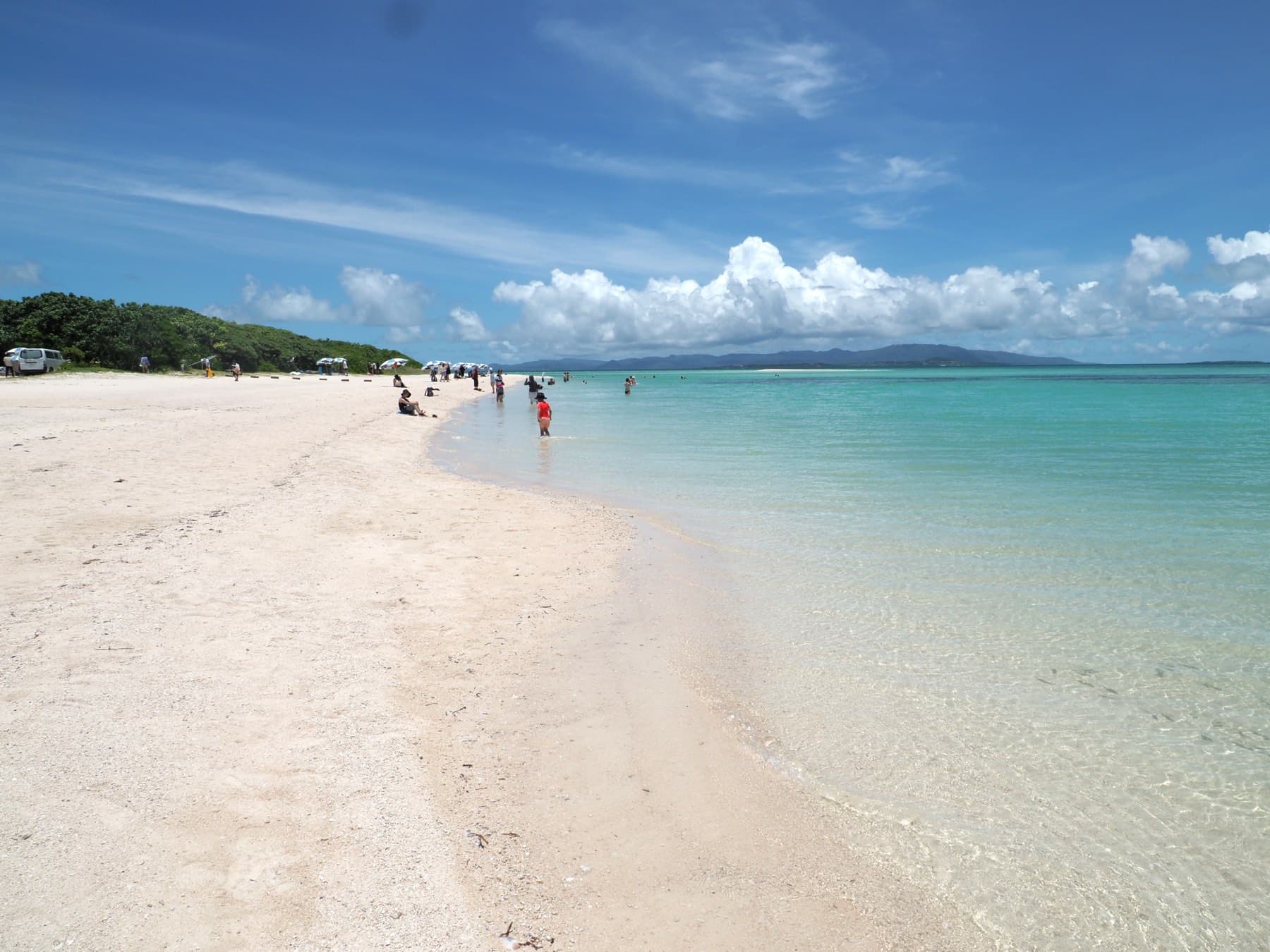 A wide band of clear green water laps the white sand of Kaiji Beach on Taketomi as a few people wade near the trees at its far end.