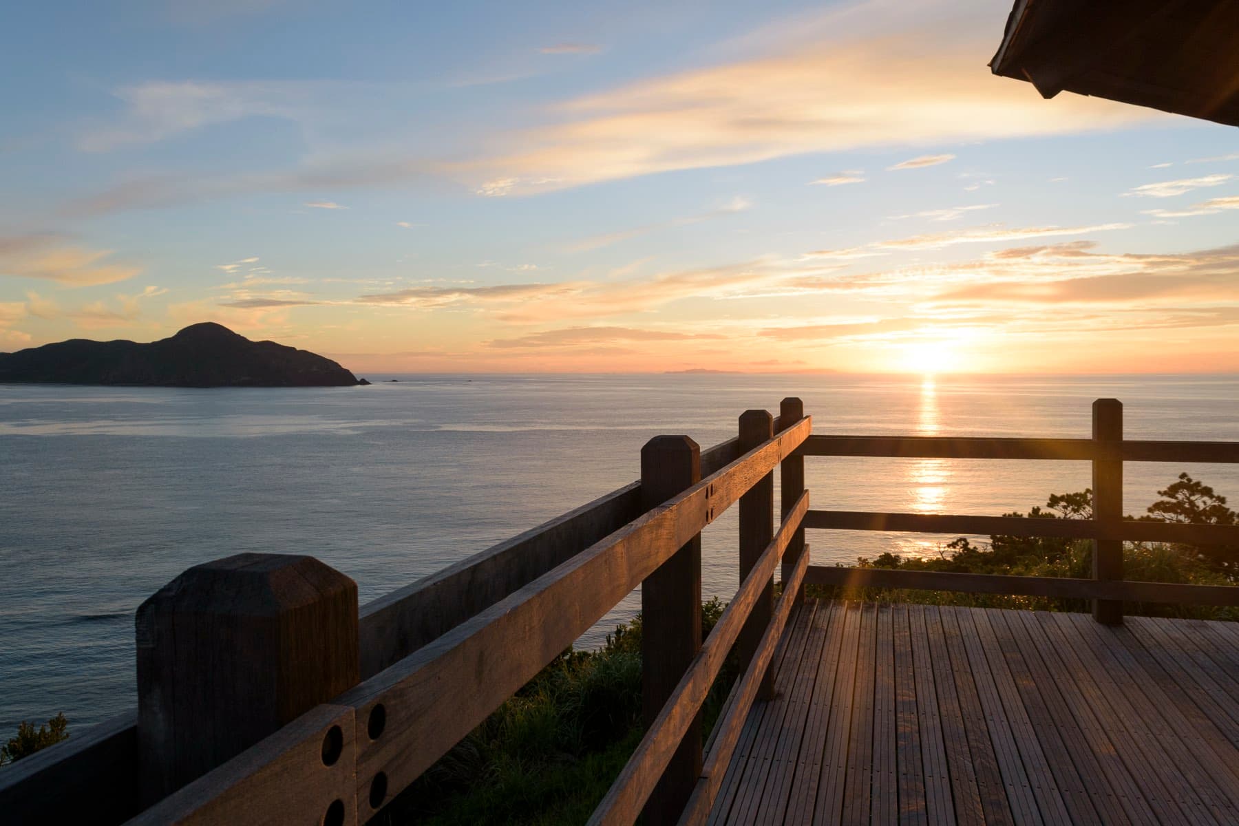 The sun drops toward a dark island on the horizon as gold light spreads across the sea below the wooden deck of the Kami-no-hama Observatory on Zamami
