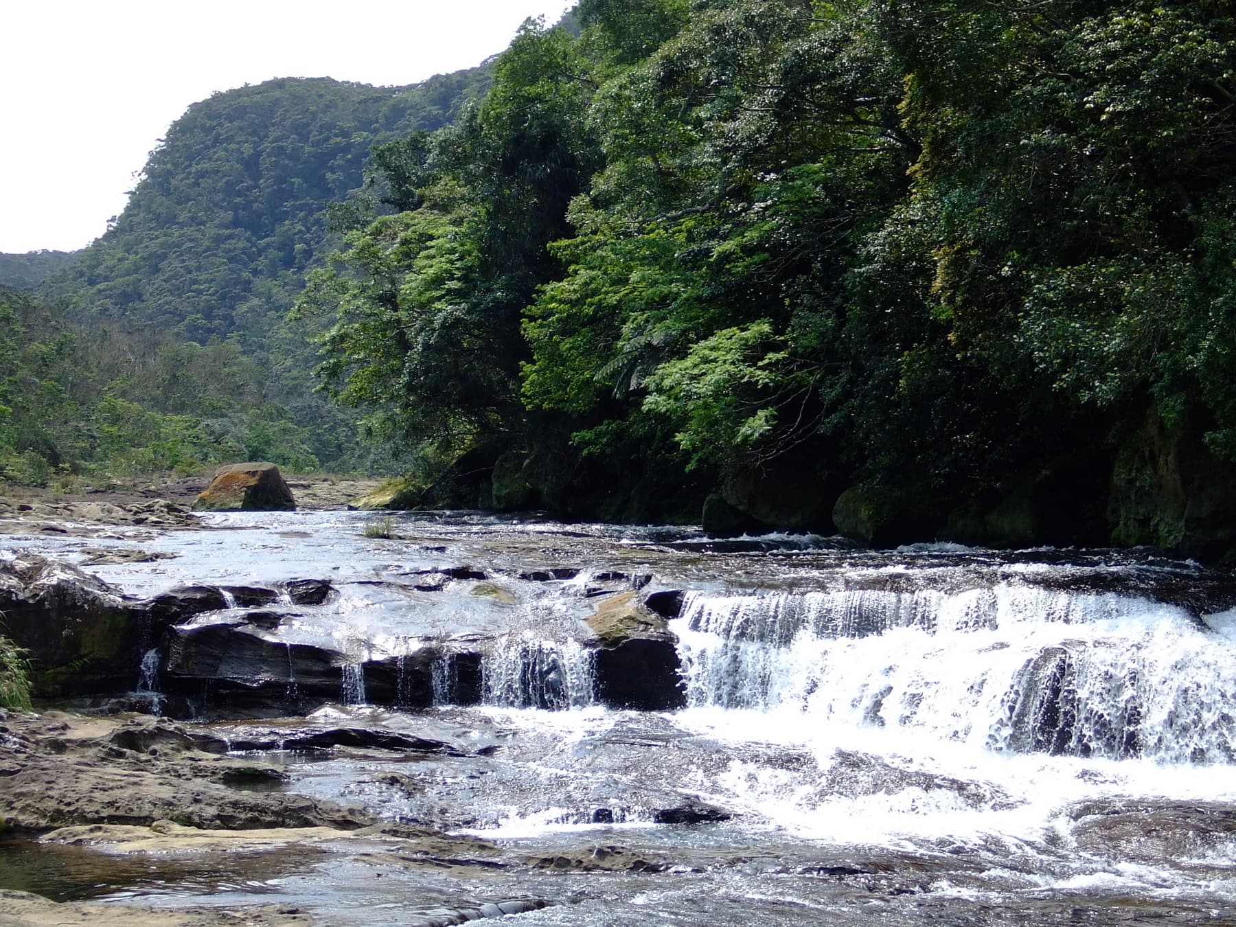White water spills over wide rock steps at Kanbire Falls on the upper Urauchi River with jungle-covered slopes on either side.