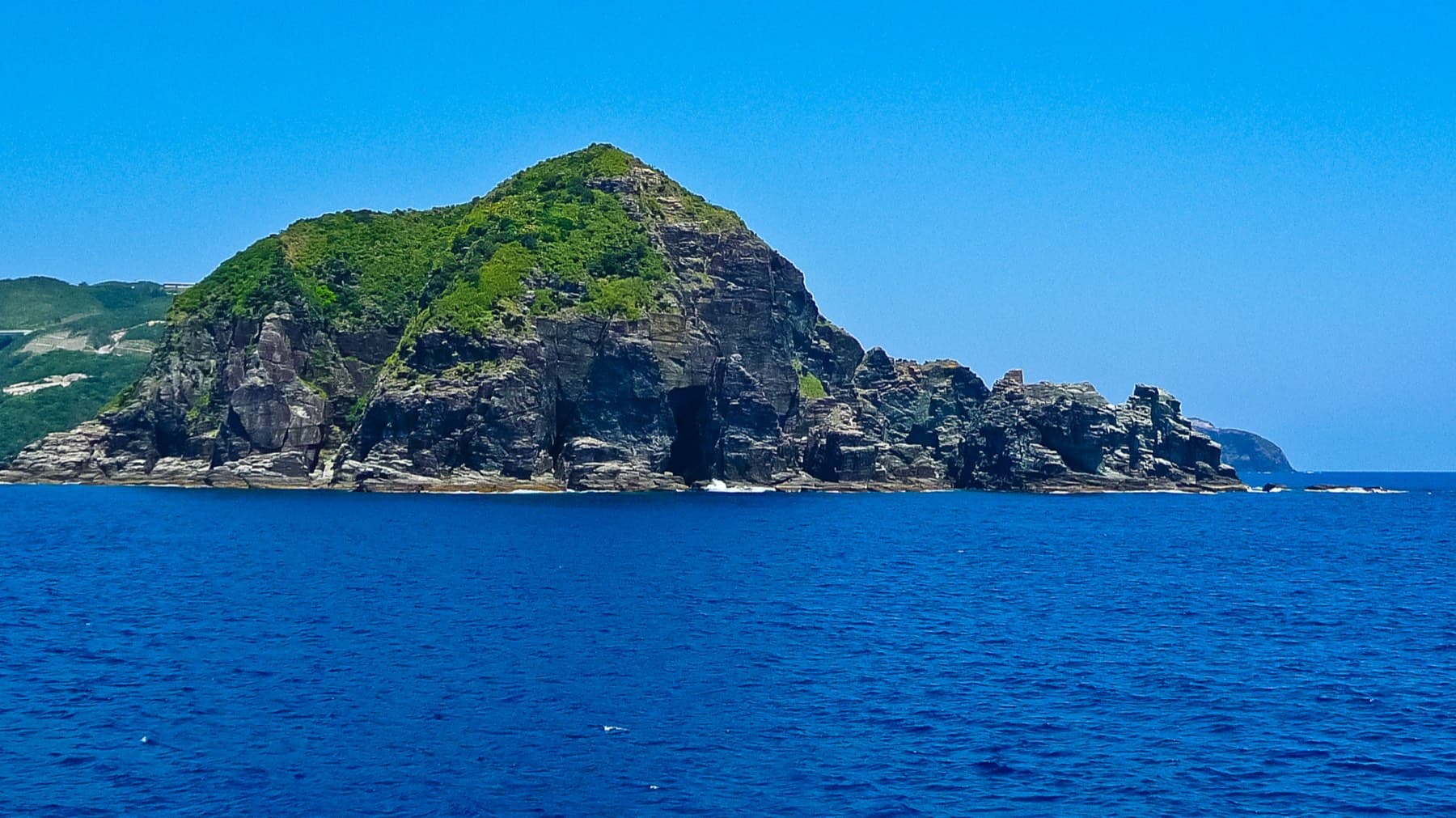 A small uninhabited islet of the Kerama group rises from deep blue water, and green scrub caps dark cliffs that open into a sea cave at the waterline