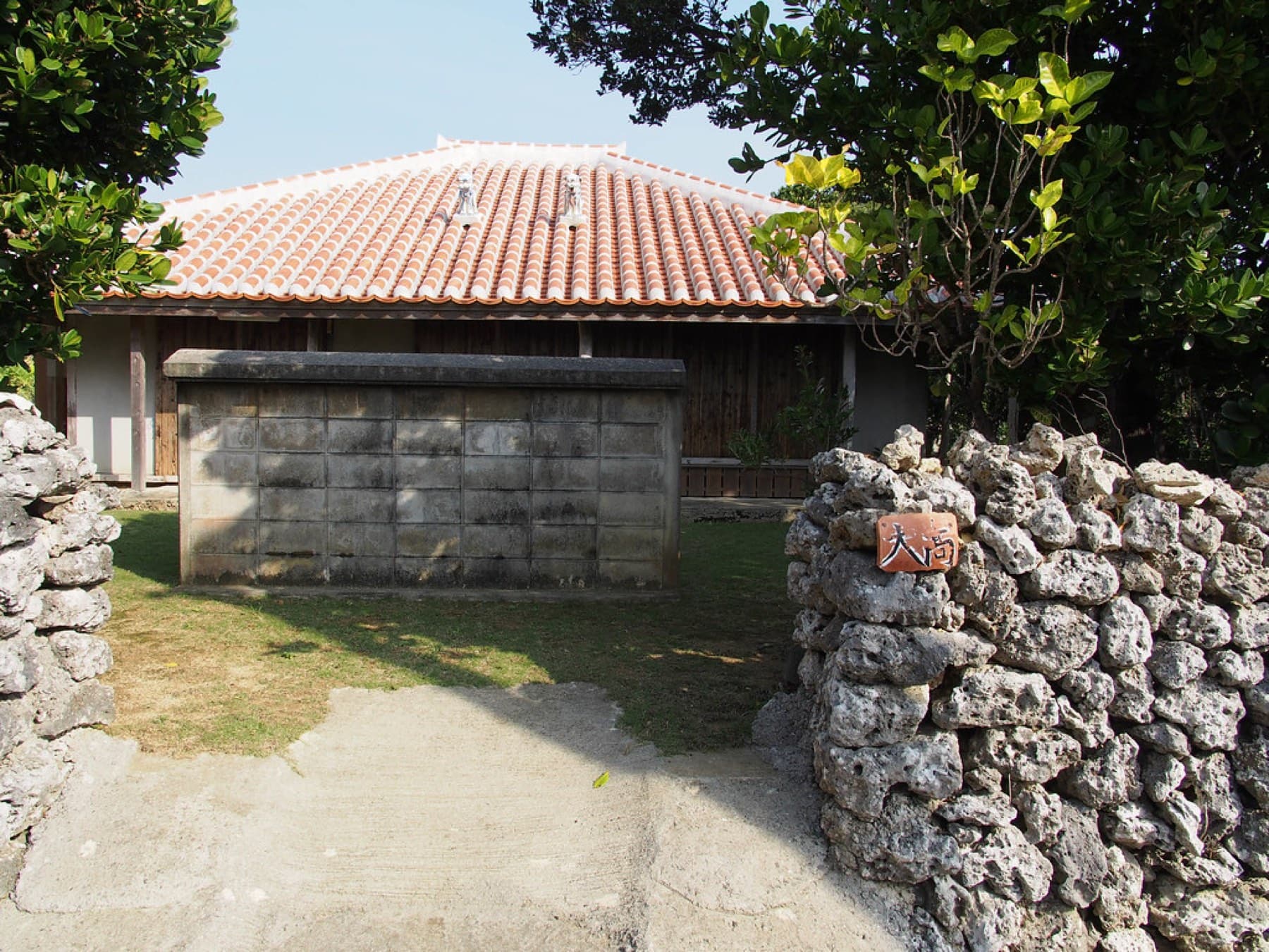 A red-tiled house stands behind a hinpun screen wall and rough coral-stone gateposts on a quiet lane in Kohama village.