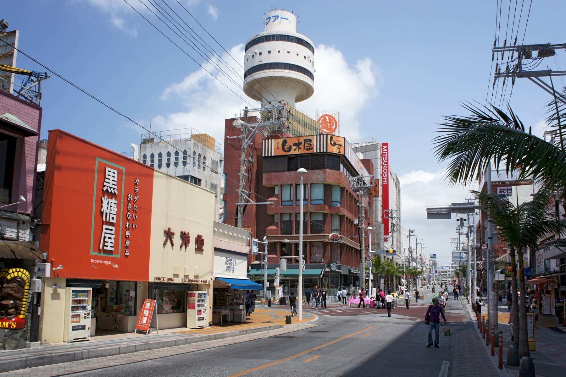 Kokusai-dori runs straight under a bright sky past a brown sugar shop and palm trees toward a round tower at the far end of the street