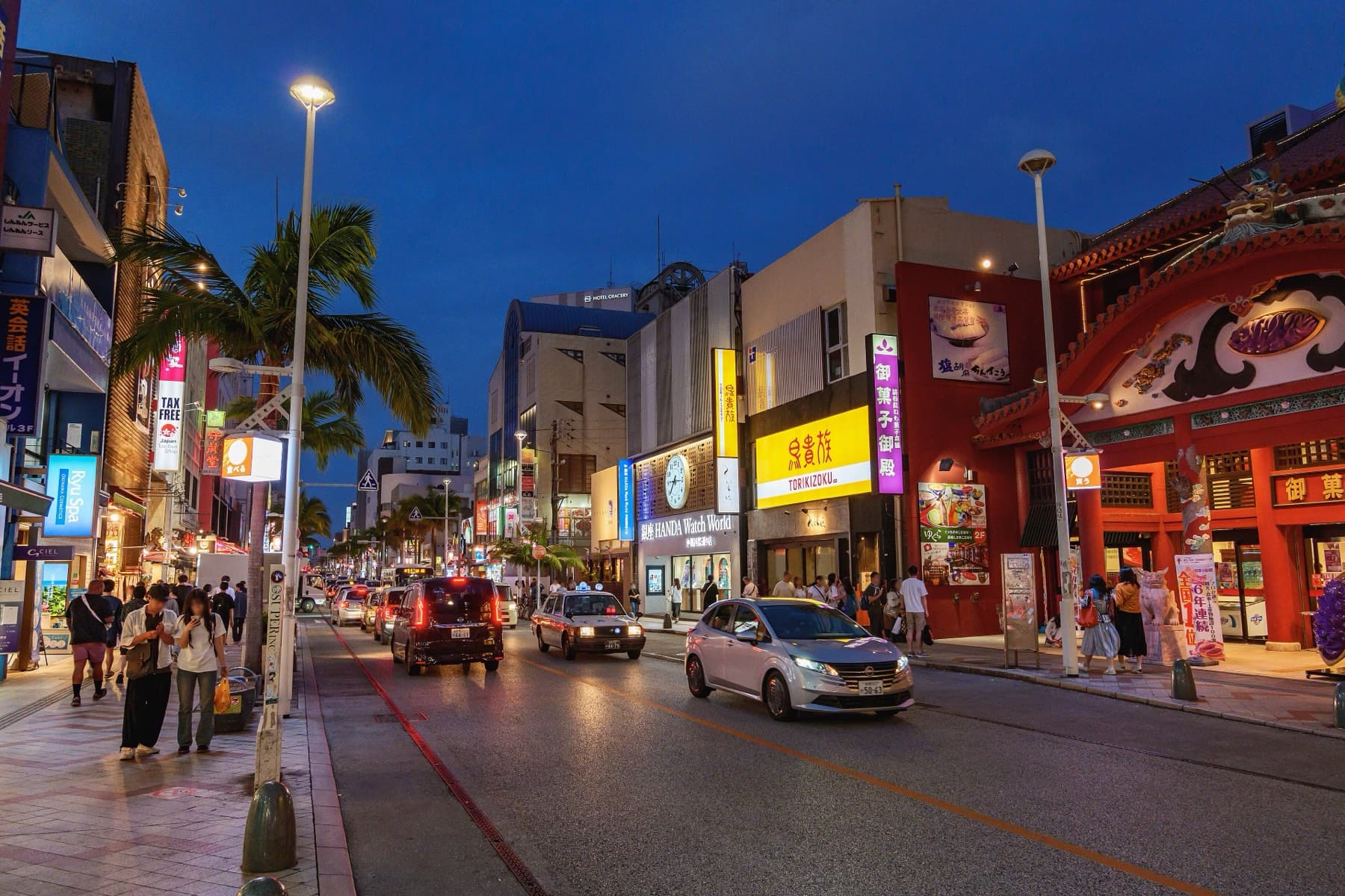 Shop signs and street lamps come on along Kokusai-dori at blue hour while taxis roll past palm trees and people walk the wide pavement