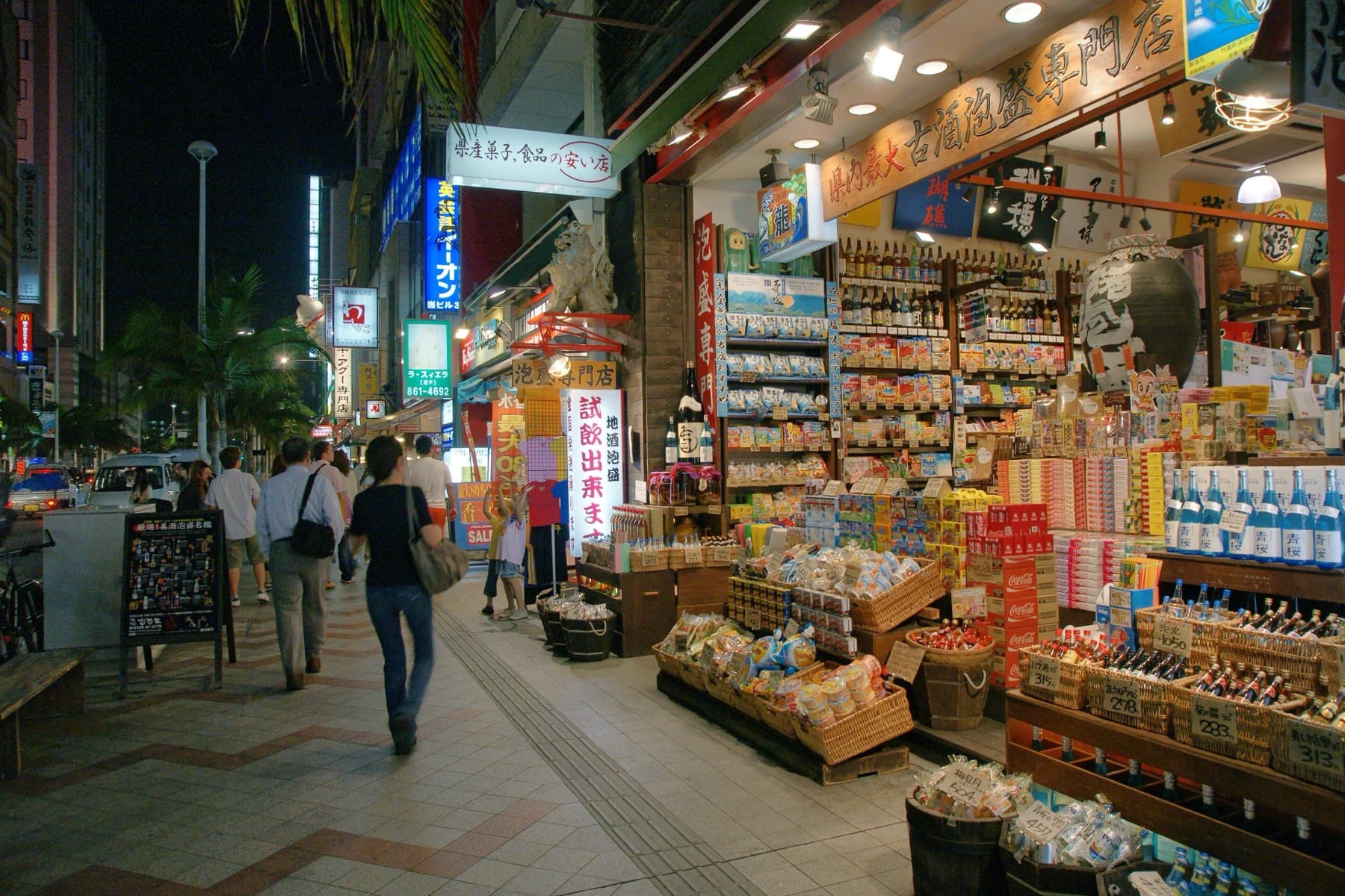 Baskets of sweets and shelves of awamori fill an open souvenir shop on Kokusai-dori at night as shoppers stroll along the arcade